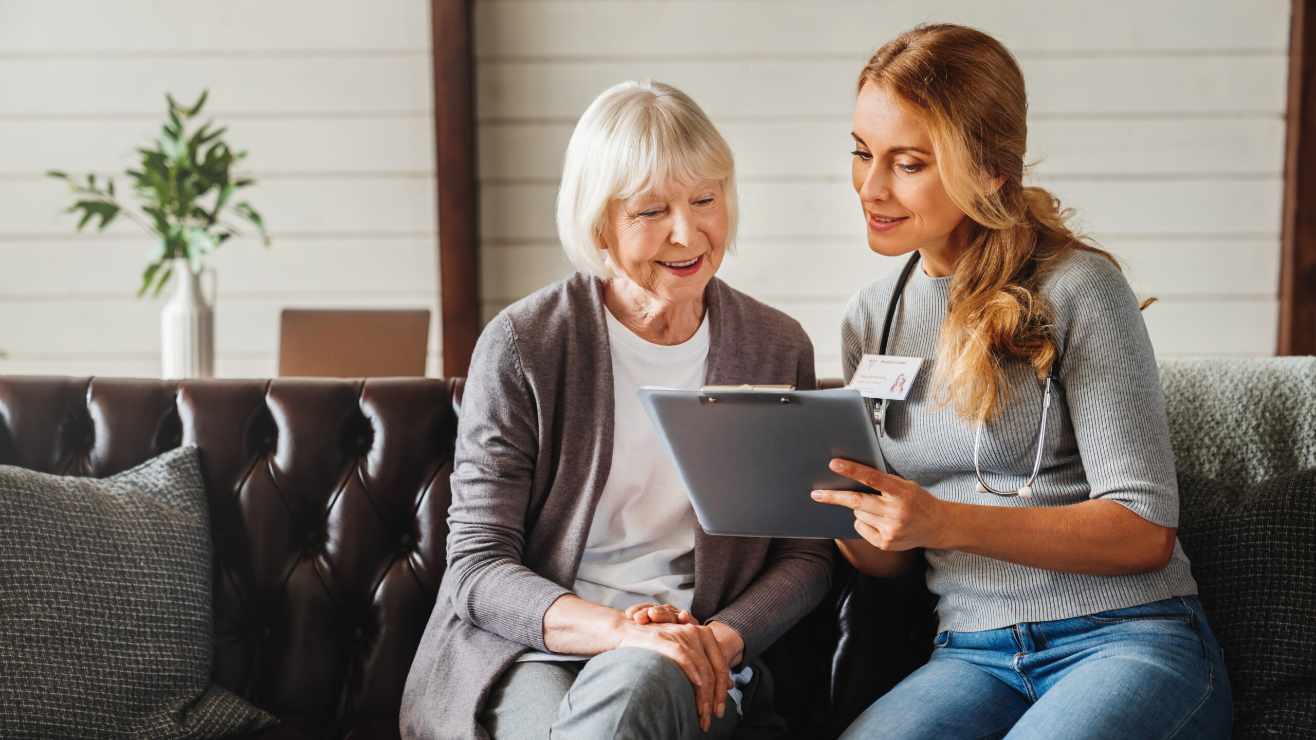 Front view shot of caring young woman nurse help old granny during homecare medical visit while making notes in clipboard. People healthcare concept